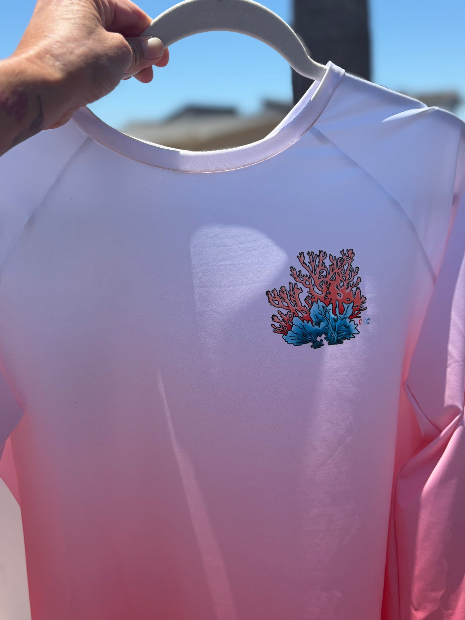 A long-sleeve coral rash guard hanging on a white hanger against a neutral wall, featuring a white-to-pink-to-orange gradient and a small coral graphic on the left chest.