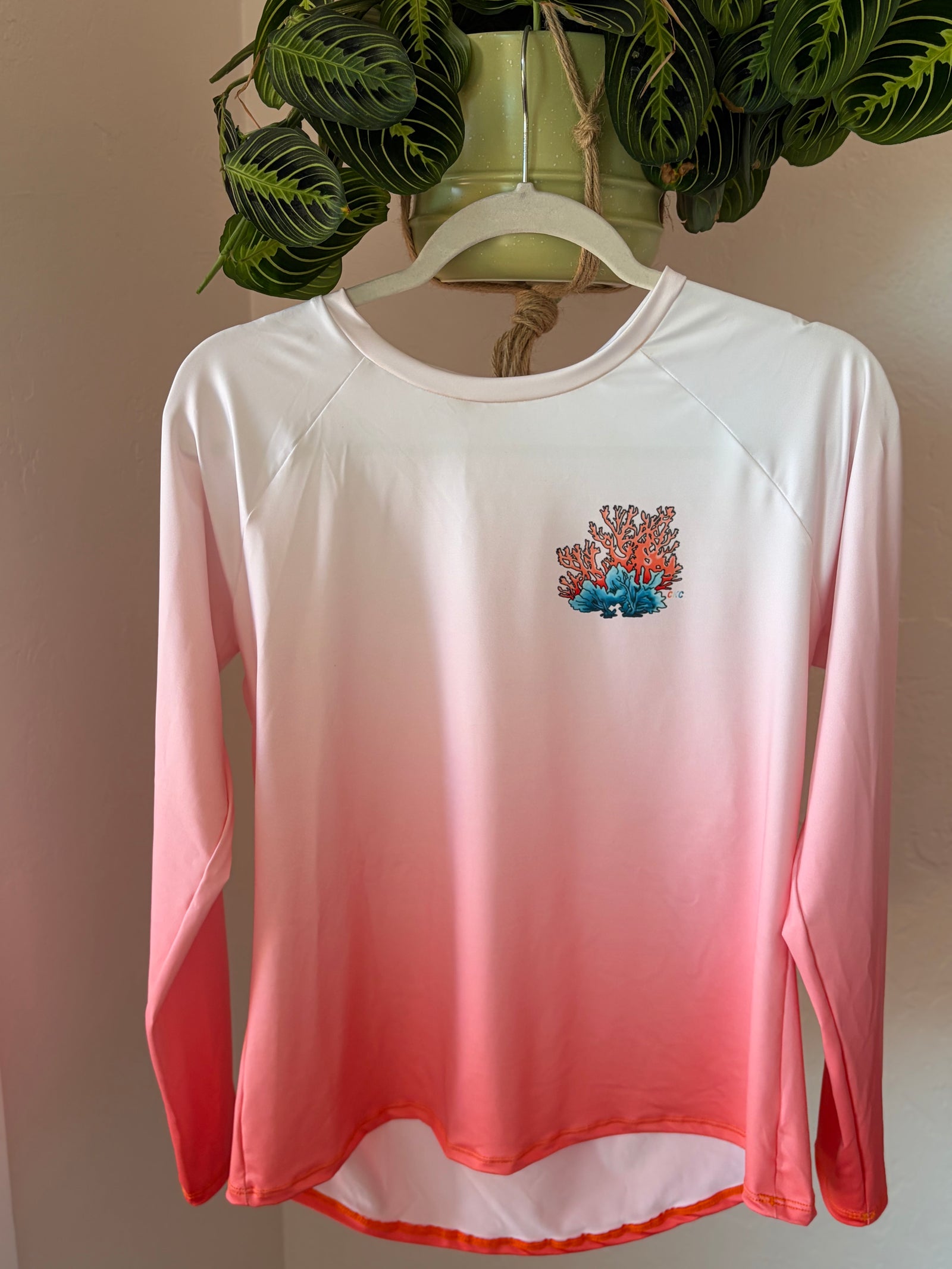 A long-sleeve coral rash guard hanging on a white hanger against a neutral wall, featuring a white-to-pink-to-orange gradient and a small coral graphic on the left chest.
