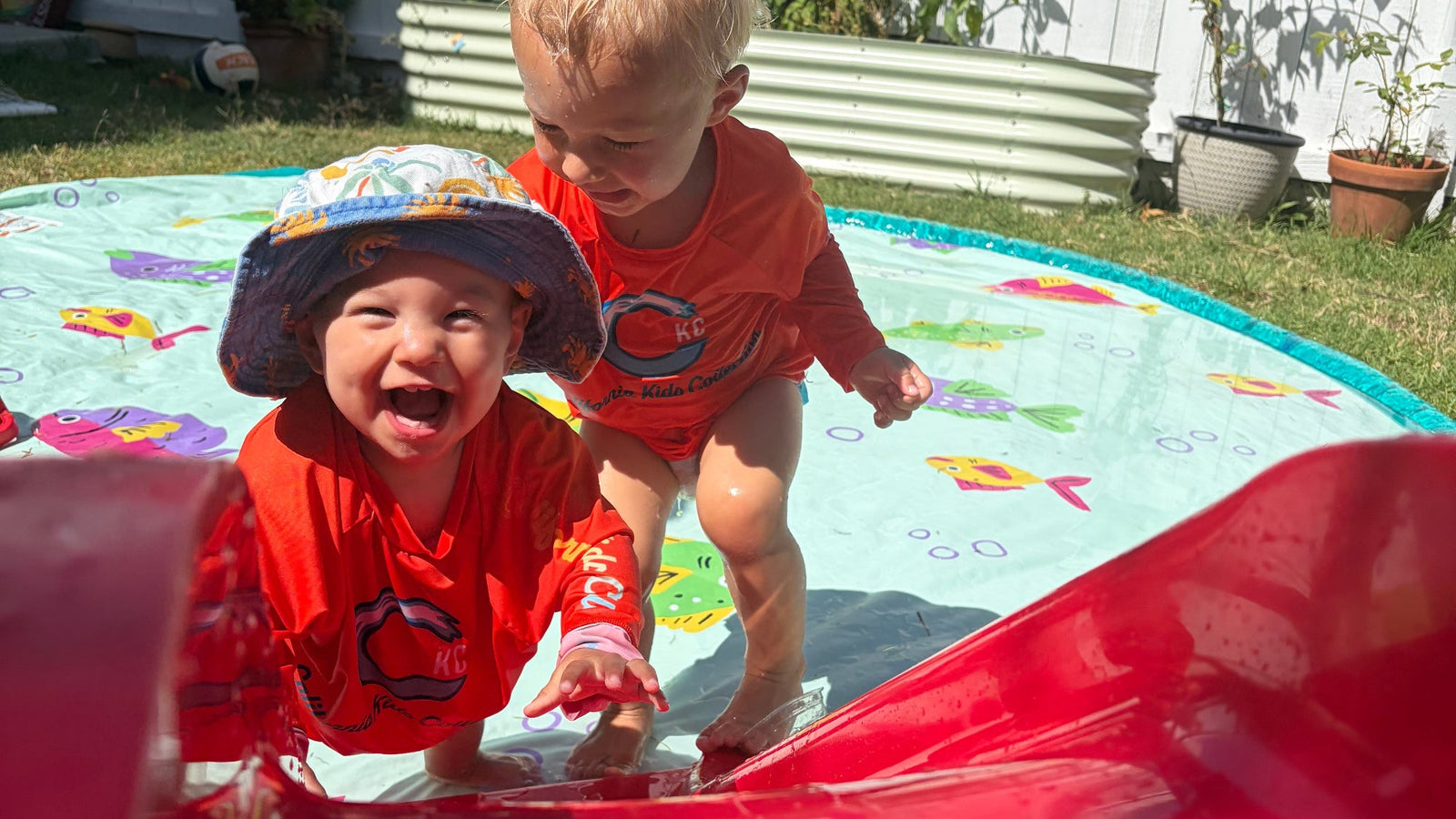 Two toddlers wearing matching red long-sleeve swim shirts play on a small outdoor pool in a backyard. One child is smiling broadly while sliding down a red slide in the foreground, and the other stands behind them on a colorful fish-patterned pool mat.