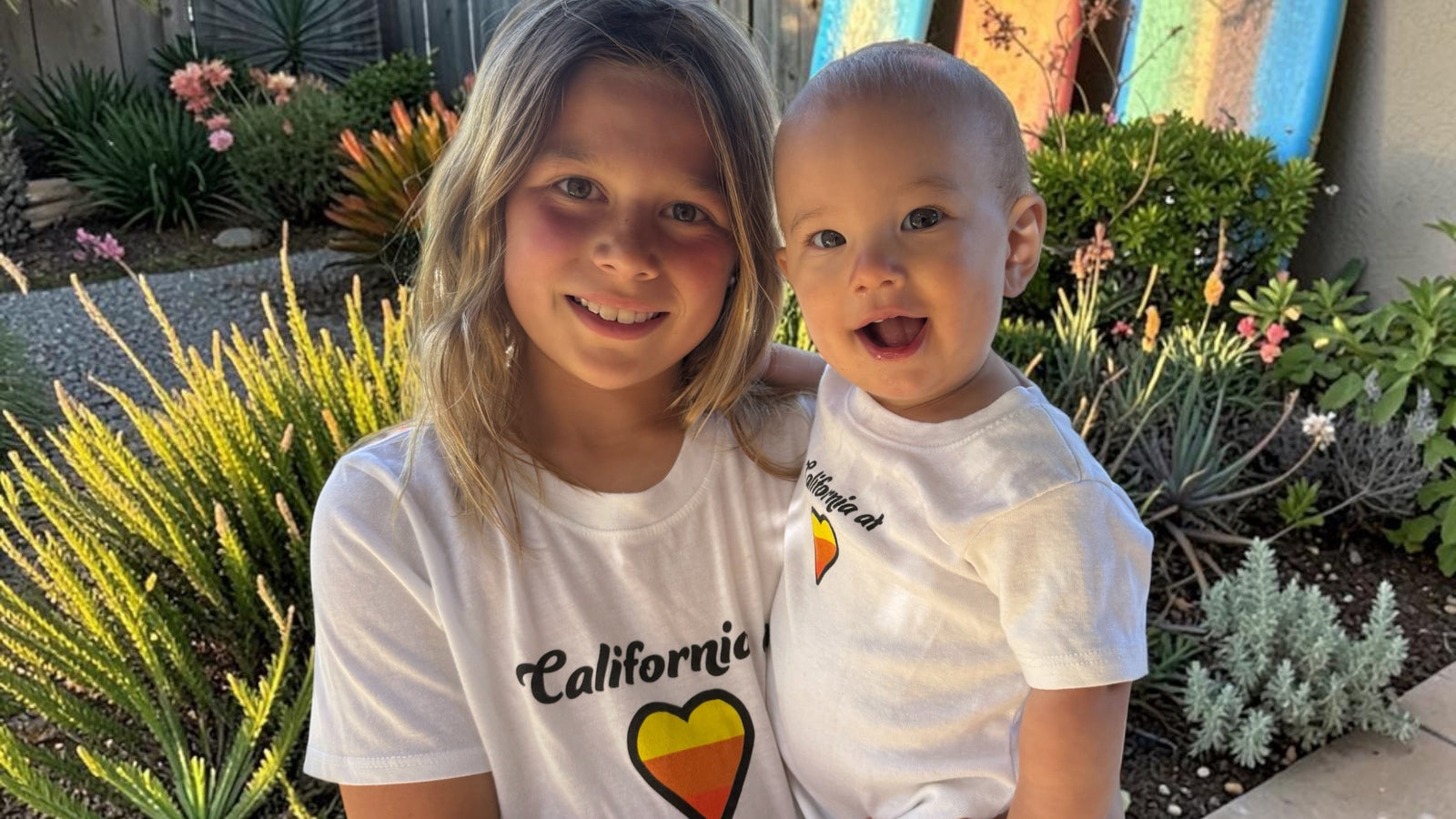Two children standing outdoors in a backyard garden setting, both wearing white t-shirts with a colorful heart graphic and the word 'California'.
