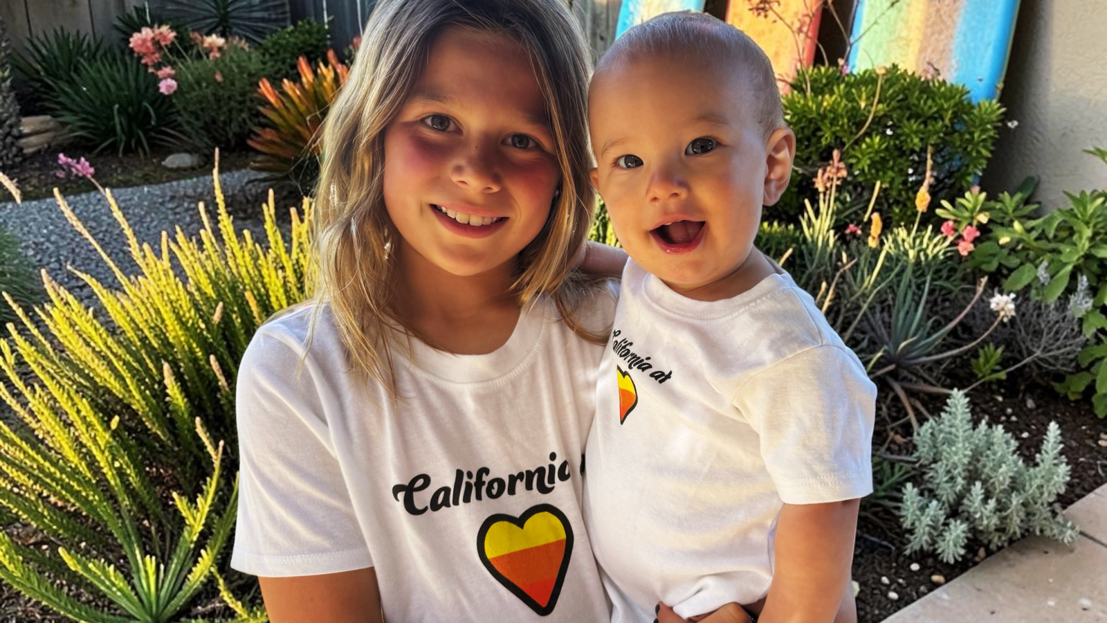Two children wearing matching white t-shirts with a 'California' heart graphic, standing outdoors in front of a garden.