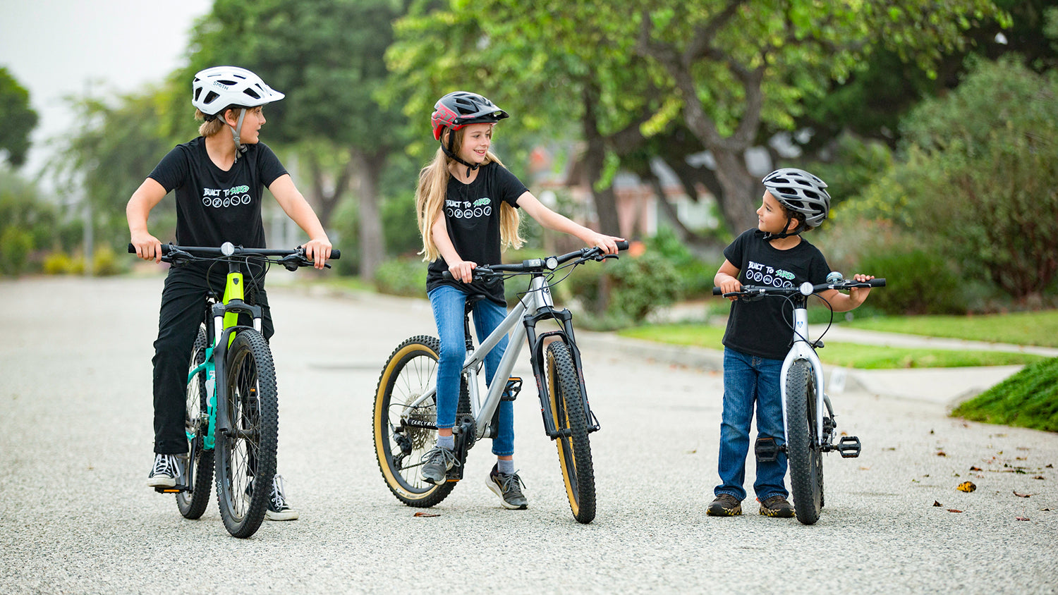 Three children wearing matching black CKC t-shirts and helmets ride or stand with bicycles on a paved path outdoors, with trees and grass in the background.