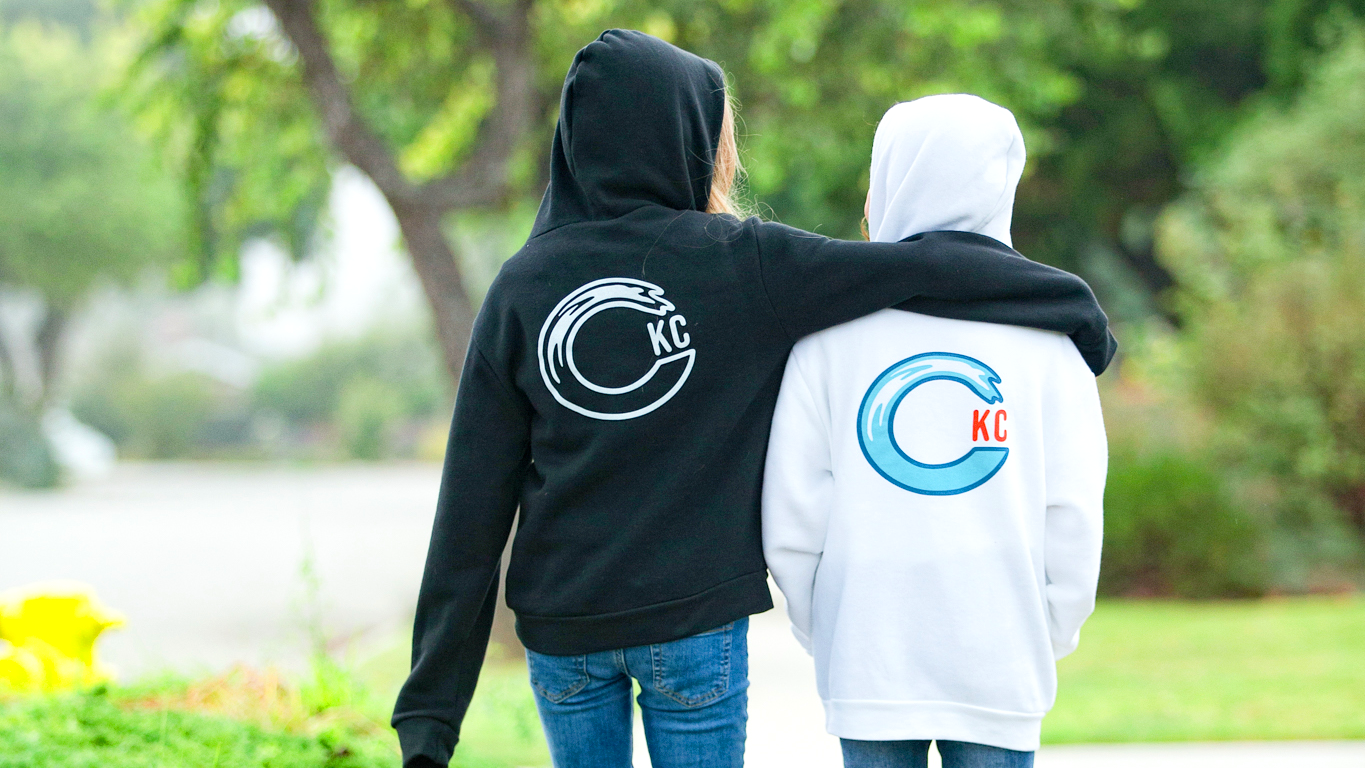 Two girls standing outdoors with their backs to the camera, wearing matching hoodies. The person on the left wears a black hoodie with a white circular CKC logo, and the person on the right wears a white hoodie with a blue circular CKC logo.